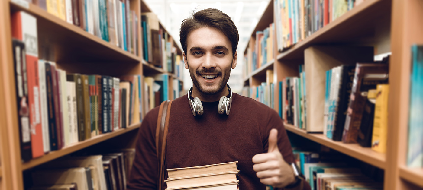 Male student in library aisle holding books and giving thumbs up sign