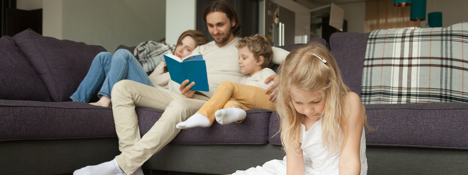 Little girl daughter playing on floor drawing while parents and boy son reading book on sofa, family spending time together in living room, weekend at home and creative child development concept