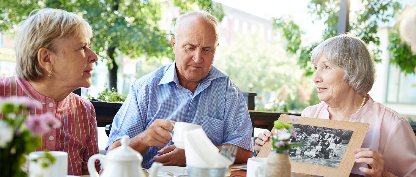 Senior friends gathered together in outdoor cafe and listening attentively to woman in pink blouse with old black-and-white photo in hands