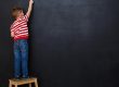 Back view of a little boy standing on ladder and writing with chalk on the backboard in school class