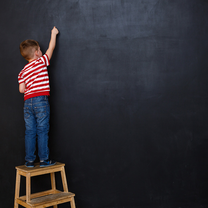 Back view of a little boy standing on ladder and writing with chalk on the backboard in school class