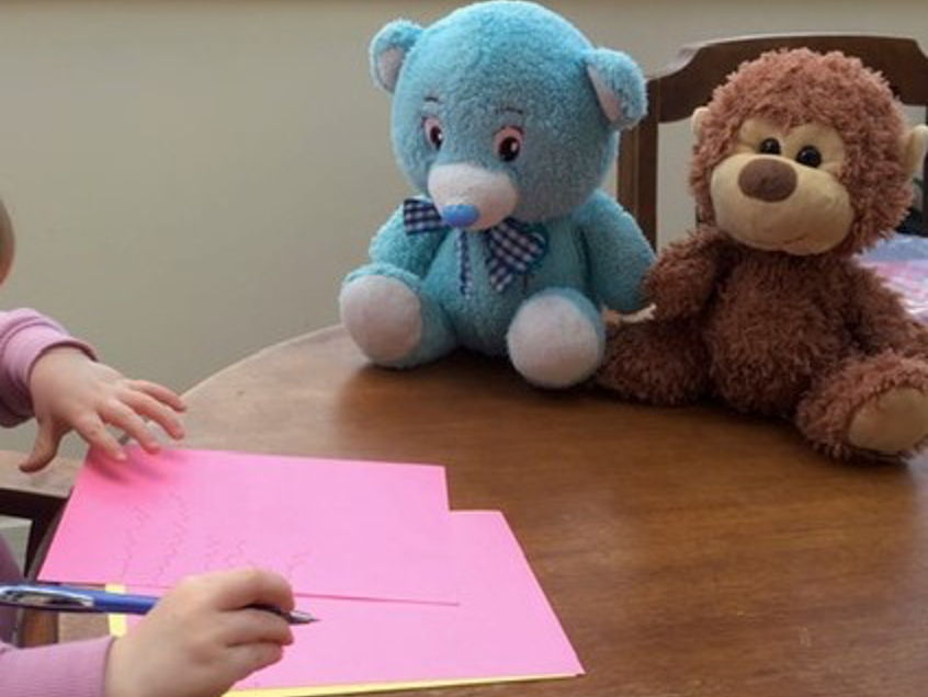 Child drawing on pink paper on desk with sort toys nearby