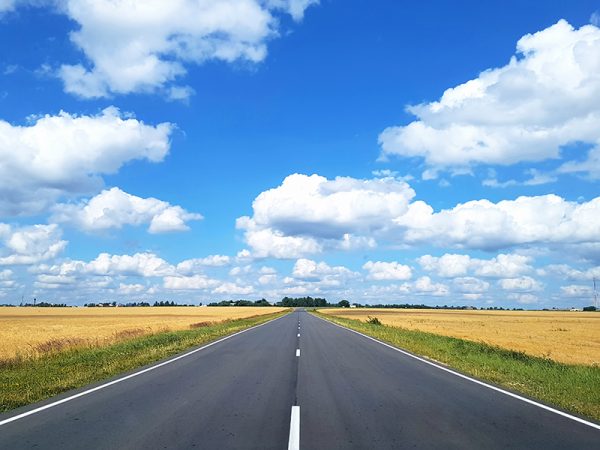 View of the asphalt highway which is in a field with wheat against the blue sky with white clouds. Concept landscape, background.