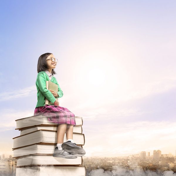 Asian cute girl with glasses holding the book while sitting on the pile of books with city and blue sky background. Back to School concept