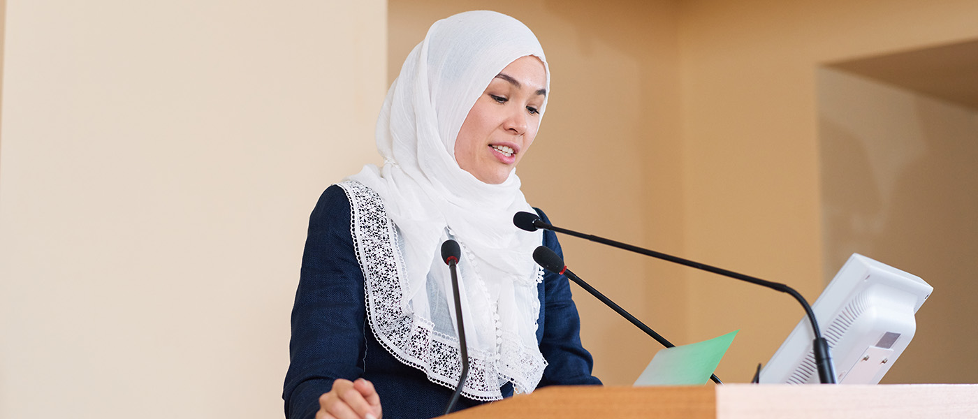 Young woman in hijab reads off screen at podium