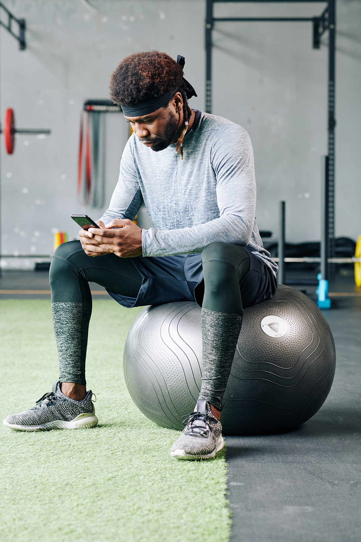 Serious young black sportsman sittting on big fitness ball in gym and reading recommendations from his coach on smartphone screen