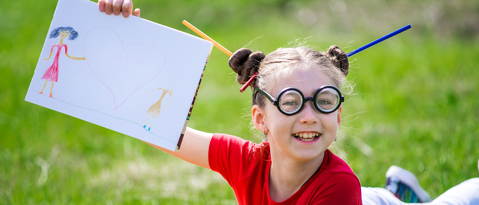 Funny girl with glasses in the park draws with a pencil in album.