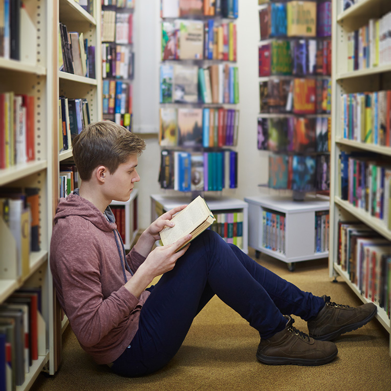 Boy taking a reading break in between library book shelves