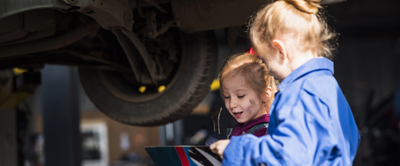Two girls with overalls holding a manual under a car on a hoist