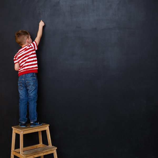 Back view of a little boy standing on ladder and writing with chalk on the backboard in school class