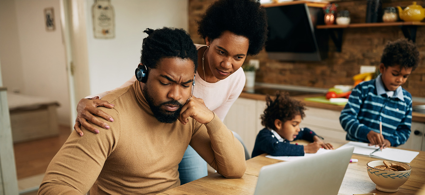 African American man writing notes and working on laptop while being at home with his wife and kids.