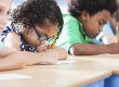 Multi-ethnic elementary school children writing in classroom. Focus on Hispanic girl wearing eyeglasses (8-9 years).