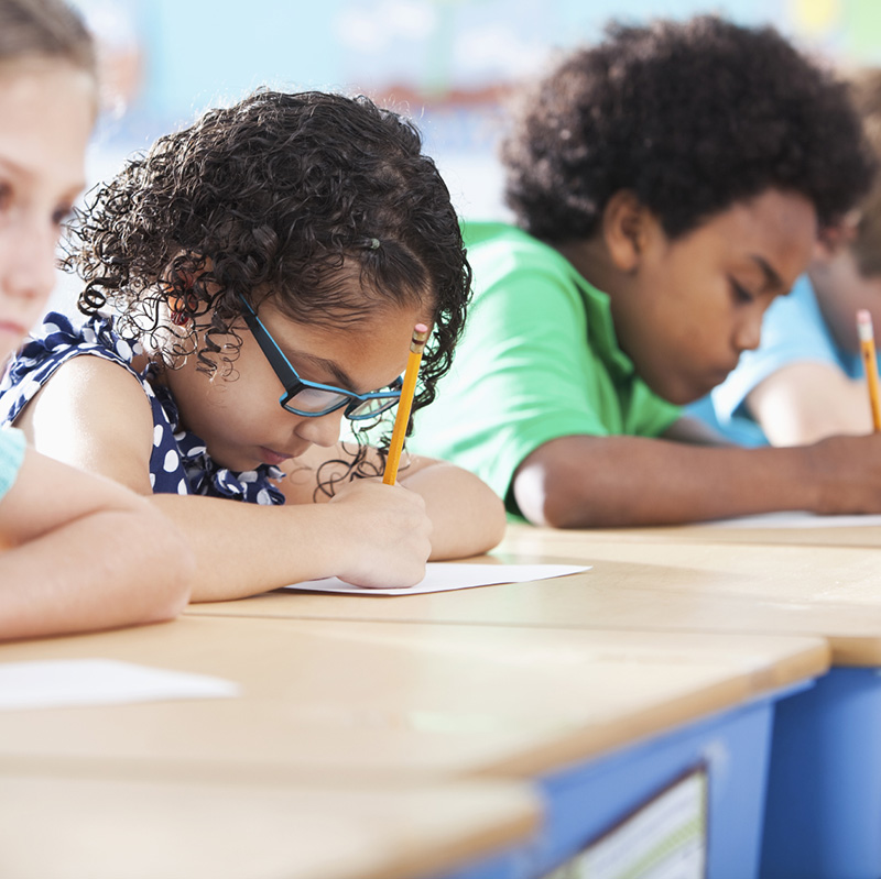 Multi-ethnic elementary school children writing in classroom. Focus on Hispanic girl wearing eyeglasses (8-9 years).